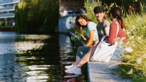 Three students sitting and talking by a river in a city.