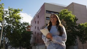 A student smilingly walks in front of the university with enrolment documents in her arms.