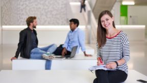 Studierende sitzt lächelnd mit Schreibblock im Foyer der Universität.