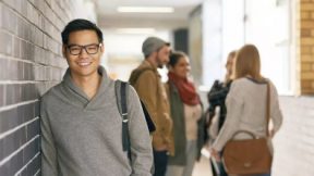 Student leans against the wall of the university building and smiles at the camera.