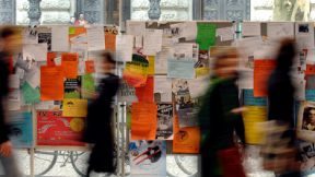 Students walk along the corridor of a university in front of the notice board.