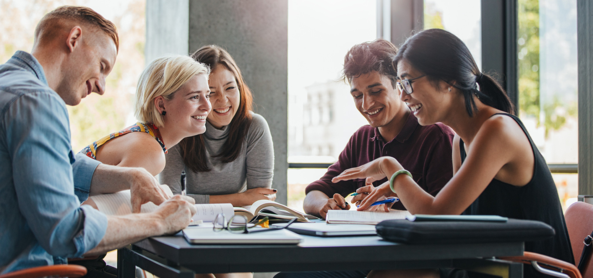 Internationale Studierende sitzen lachend an einem Tisch in der Universität und lernen gemeinsam.